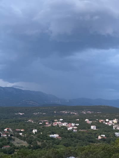 This media file depicts a twilight scene in Višnjevo, Montenegro, focusing on a hillside dotted with houses nestled amongst lush greenery. The sky is predominantly a deep, moody blue, filled with heavy, dark clouds that suggest impending rain or a recent storm. Faint hints of lighter clouds can be seen, creating a textured, atmospheric effect.

To the right of the frame, the steep, tree-covered mountainside dominates. Scattered across its slope are numerous buildings, ranging from small houses to larger villas, their roofs a mix of red, white, and other muted tones. Many of these homes are illuminated, with small points of light visible, indicating that it is evening. The dense foliage of the trees creates a thick carpet over the mountain, with some clearer patches revealing the terrain beneath.

In the background, further mountain ranges are visible, shrouded in a hazy, bluish light that emphasizes their distance and the deepening dusk. The overall impression is one of a serene, somewhat isolated community set against a dramatic natural backdrop. The presence of clouds and the subdued lighting suggest a late afternoon or early evening, possibly with the threat of rain adding a dramatic element to the otherwise peaceful landscape. No people or specific activities are discernible. The image is oriented vertically, with the sky occupying the left side and the mountainside the right.