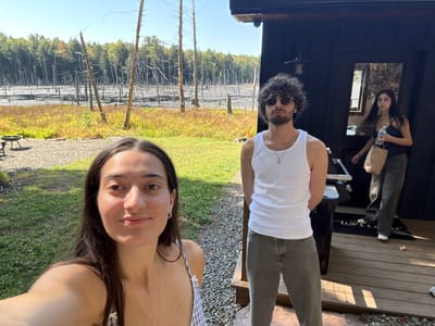 The scene is set in a rural location near Otego, United States, featuring three individuals outdoors on a clear day. In the immediate foreground, a female with long, dark hair, wearing a striped or plaid strappy top, looks directly at the camera with a slight smile. Centered in the middle ground, a male with dark curly hair, a beard, and sunglasses stands with hands clasped behind his back, clad in a white tank top and light-colored pants. To the right, a second female exits a dark-colored cabin-style building, holding a clear plastic bottle in her right hand and a brown paper bag in her left, dressed in a dark top and light wide-leg pants. The background reveals a marshland or flooded forest containing numerous bare, dead trees (snags) standing in water, bordered by a dense green forest under a blue sky. The ground in the foreground consists of grass and gravel, with a metal fire pit located on the far left. The building entrance features a "Welcome" mat and an external light fixture.