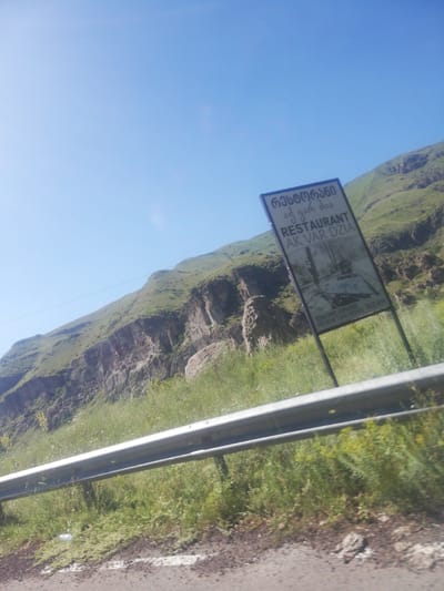 The image shows a scenic view of a mountainous landscape in Gogasheni, Georgia. A roadside sign is prominently displayed, advertising a restaurant. The sign is black and white and contains text in both Georgian and English. The Georgian text reads "რესტორანი" which translates to "Restaurant". Below this, in English, it says "RESTAURANT AK VAR DZIA". Further down the sign, there is smaller text that appears to be a phone number and a mention of "MESKHETIAN DISHES 1500m". The mountainous terrain is covered in green grass and some rocky outcrops. The sky is a clear blue. In the foreground, there is a metal guardrail along the side of what appears to be a road, with more greenery visible behind it. The overall impression is of a natural, rural setting, likely a tourist destination.