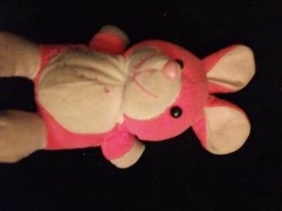 A pink plush toy lies against a dark background. It appears worn with some discoloration and dirt visible on its beige sections. The toy features a round pink nose and black button eyes. Its ears are rounded and have a beige interior. The toy's body appears stuffed with visible stitching. The overall image has a slightly blurred quality.
