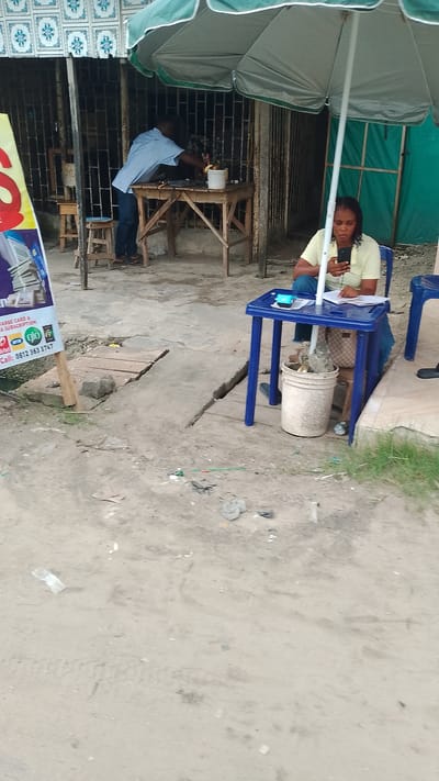 The image captures a street scene in Agbarho, Nigeria, likely a commercial area given the outdoor setup. In the foreground, a woman sits at a small blue plastic table under a green and white umbrella, looking at her mobile phone. The table has papers and a small blue object on it, and a white bucket is placed beneath it. Beside her, another blue plastic chair is visible.
Behind the woman, a man in a checkered blue and white shirt and dark pants is standing at a wooden table, appearing to be working or attending to something. A smaller wooden stool is nearby. The background features a building with barred windows and a patterned tiled facade. Various debris and some grassy patches are visible on the ground, suggesting an unpaved or worn-down urban environment. A partially visible advertisement poster is also present on the left side of the image. The overall impression is of an outdoor business or service point on a street.