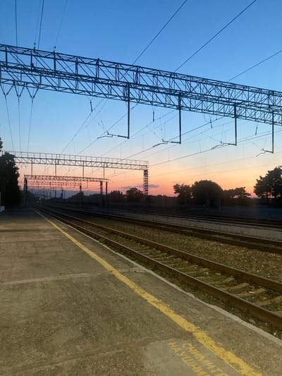The image depicts a railway station at dusk in Samur, Russia. In the foreground, a concrete platform with a yellow safety line is visible, bordering multiple sets of train tracks. Overhead, a complex network of power lines and supporting structures for an electric railway is silhouetted against a gradient sky transitioning from deep blue to pale orange and pink. Trees and distant hills form the horizon line. The tracks appear to be in use, indicated by their presence and the overhead electrification infrastructure. There are no people visible in the scene. The overall atmosphere is serene and indicative of the end of the day.