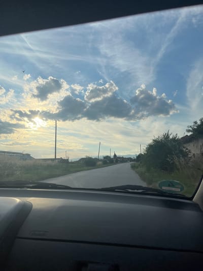 This is a forward-facing, medium-angle, eye-level, full-shot photograph taken from inside a vehicle, looking out through the windshield. The scene depicts a rural road with power lines and scattered buildings under a cloudy sky with the sun breaking through. The road appears paved and relatively narrow, curving slightly into the distance. On the left side of the road, there are fields and a large, long building. On the right side, there are trees, bushes, and the side of a weathered building. The sky is a mix of blue and white clouds, with the sun creating bright rays. A single bird is visible in flight in the upper left portion of the sky. A green circular sticker with white markings is affixed to the interior of the windshield on the right side. The interior of the vehicle, specifically the dashboard, is visible in the foreground. The location is identified as the city of Novo Zhelezare, Bulgaria.