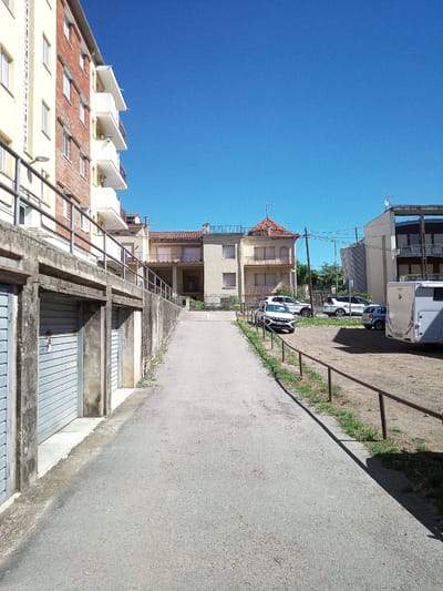 The image shows a sunny day in the town of Castellfollit de la Roca, Spain, with a clear blue sky. In the foreground, there is a concrete walkway or ramp leading uphill, bordered by a metal railing. To the left of the walkway are several garage doors with a corrugated metal texture. Further up the walkway, on the right, is a gravel parking area with several cars parked: a white SUV, a dark SUV, a small blue car, and a white camper van. Behind the parking area are several buildings, including a light-colored apartment building with red brick accents and balconies, and another building with a darker facade and balconies. The overall scene depicts a residential area with attached garages and outdoor parking under a bright sky.