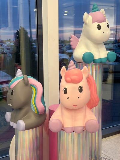 The image displays three cartoon-style unicorn figures arranged vertically against a reflective surface. The topmost unicorn is predominantly white with a light purple mane and teal details. Below it is a pink unicorn with a coral mane. The lowest unicorn is gray with a multicolored mane of pink, teal, yellow, and white. The unicorns are set in what appears to be a store window based on the reflections and the objects partially visible in the background. It is impossible to verify that this scene is in the city of Novoaleksandrovka, Russia.