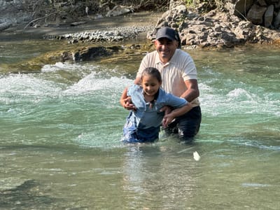 A man and a young girl stand in a shallow, flowing river. The man, wearing a light-colored polo shirt and a dark baseball cap, has his arms around the girl. The girl, who appears to be wearing a denim shirt and dark pants, is looking down and smiling. The river water is clear and green, with some ripples and small waves. The background shows a rocky, vegetated bank. The overall scene depicts a familial moment in a natural outdoor setting.