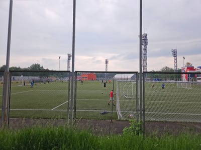 A view of a football field in Sosnovoborsk, Russia, through a chain-link fence. Several people, some appearing to be playing football, are visible on the green artificial turf. Several tall light towers are positioned around the field. In the background, there are buildings, trees, and a cloudy sky. The foreground is partially obscured by tall green grass.