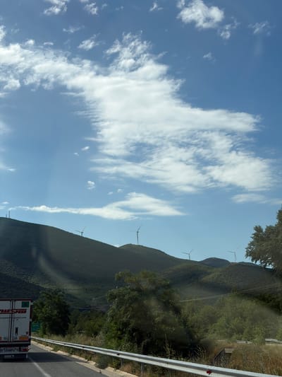 This is a medium shot, captured from a vehicle, looking out at a rural landscape under a partly cloudy sky. In the foreground, the right side of a white truck with red and black lettering is visible. The truck has a license plate that reads "FKG 6503 SCHMITZ". To the right of the truck, a road curves away, bordered by a silver guardrail. Beyond the road, a dense line of green trees and vegetation obscures the ground, with glimpses of power lines stretching across. The mid-ground is dominated by rolling, tree-covered hills. Several wind turbines, with their blades extended, are positioned on the crests of these hills. In the distance, the sky is a bright blue, with scattered white clouds. The scene appears to be set in a rural area of Δημοτική Ενότητα Προμαχώνος, Greece, likely a highway passing through a hilly region with wind farms.