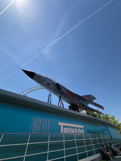This media file captures a dramatic upward-angled shot of a roller coaster, a simulated jet fighter, and a clear blue sky. The jet fighter, painted in shades of gray with red accents and the number "3057" visible on its side, is the primary object, tilted as if in flight or descending. It's mounted on a roller coaster track, suggesting it's part of an amusement park ride.

On the right side of the frame, a light blue building or structure with the word "TORNADO" prominently displayed in large, stylized white letters dominates the space. Below "TORNADO," the word "BLUE" is also visible, also in large, stylized letters. This building appears to be part of the roller coaster's infrastructure, possibly its station or a themed element.

Several people are visible in the upper right section, seated in what appear to be roller coaster cars or a viewing area. They are positioned on the blue structure, looking towards the jet fighter or the ride's path. Their posture suggests they are either waiting for the ride or enjoying it.

The sky is a bright, cloudless blue, with the sun casting a strong light towards the bottom left, creating lens flare. Two distinct contrails are visible across the sky, one sharp and linear, the other more diffuse, hinting at air traffic. The presence of trees can be seen at the very top right, suggesting an outdoor setting. The overall atmosphere is bright and sunny, indicative of a clear day. The setting is likely an amusement park, given the roller coaster and themed elements. The visible text "TORNADO" and "BLUE" strongly suggest the name of the ride or park attraction. The location context of Castelnuovo del Garda, Italy, would place this at a specific amusement park in that region.