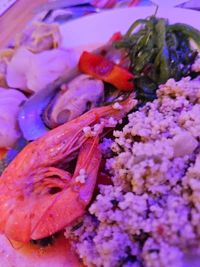 A close-up, high-angle image depicts a prepared meal served on a white plate in Audun-le-Tiche, France. The scene is illuminated by artificial lighting, casting distinct purple and pink hues across the food items. Prominently in the foreground are two cooked, head-on shrimp with orange-pink exoskeletons; one shrimp displays small white particulate matter on its shell. To the immediate left, two mussels are visible, one within a blue-black shell and another lighter-toned, partially open. A large mound of granular food, consistent with couscous or a similar grain salad containing discernible green and red flecks, occupies the right side of the plate. Above this, a portion of dark green, stringy seaweed salad is present, adjacent to a single red bell pepper slice. In the upper left background, several light-colored, folded items resembling dumplings or wontons are partially obscured. No individuals are visible.