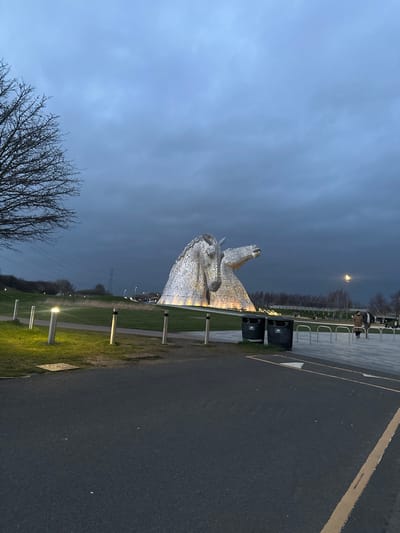 The image depicts an exterior scene at dusk or early evening at The Kelpies, Helix Park, Falkirk, United Kingdom, under a heavily overcast, dark grey sky. The primary subjects are two monumental, illuminated metallic horse-head sculptures, appearing silver, positioned in the mid-ground. The left sculpture faces slightly left, while the right sculpture faces right with its mouth open. Both are mounted on a partially lit base.
The foreground features a dark grey asphalt surface, with a single yellow dashed line along its right edge. To the left, a paved pathway is bordered by a series of short, cylindrical, illuminated bollard lights casting a warm glow onto the adjacent grass. Two dark waste bins are situated on a lighter paved area to the right of the pathway, immediately in front of a metallic bike rack.
In the upper left, a large, bare-branched deciduous tree stands against the sky. The mid-ground and background beyond the foreground paving consist of an expansive area of green and brown grass. In the distant left background, a large electricity pylon and associated power lines are visible. Further along the horizon, a tree line and indistinct road infrastructure with faint vehicle lights are present.
Two individuals are visible in the far right mid-ground, walking away from the viewer on a paved surface near the base of the sculptures. One person wears a light-colored coat; the second individual is less clearly defined. No direct interactions with the sculptures are observable.