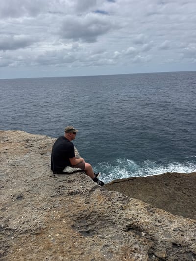 A person sits on the edge of a cliff overlooking the ocean. The person wears a dark shirt, light-colored shorts, shoes, and a hat, gazing out at the water. The cliff face is rocky and textured. The sea is dark and turbulent, with waves crashing against the cliff base. The sky is overcast with low-hanging clouds.