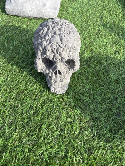 The image depicts a gray, skull-shaped object resting on artificial green turf. A rectangular gray object is partially visible on the left edge of the frame. The environment appears to be outdoors. The location is Chirk, United Kingdom.