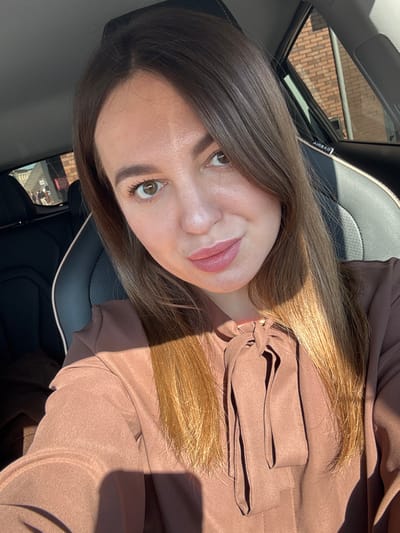 An adult female with light brown hair and light brown eyes, wearing a brown long-sleeved blouse with a pussy-bow necktie, is depicted in a selfie-style photograph. She is seated in the front passenger seat of a vehicle, identifiable by black and light-colored leather upholstery and an "AIRBAG" label visible on the pillar above the passenger seat. Strong sunlight enters from the right, casting distinct shadows on her hair, right cheek, and the front of her blouse. Through the rear passenger window on the left, an exterior scene is visible, showing a reddish-brown brick building and several indistinct figures of individuals walking. The scene is located in Likino-Dulyovo, Russia.