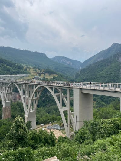 This media file captures a scenic view of the Đurđevića Tara Bridge in Montenegro, a large concrete arch bridge spanning a dramatic river canyon. The image is oriented vertically, with the sky at the top and the canyon floor at the bottom.
The primary subject is the bridge itself, a massive concrete structure with multiple arches and supporting columns. The bridge is lined with people, appearing as small figures on the walkway, suggesting it is open to pedestrians or tourists. Some vehicles are also visible on the bridge's roadway. Surrounding the bridge are lush, green mountainsides covered in dense forest, indicative of a natural, possibly rural, setting. The sky is overcast with clouds, suggesting a diffused light and potentially a moderate temperature.
In the distance, a small settlement or village can be seen nestled on a hillside amongst the trees. There are piles of construction materials visible near the base of the bridge, hinting at ongoing work or maintenance. The overall impression is one of impressive human engineering set against a powerful natural landscape. The activity appears to be the normal functioning of a bridge, with people traversing it and enjoying the views. The time of day is not explicitly discernible but the overcast sky suggests it is daytime.
