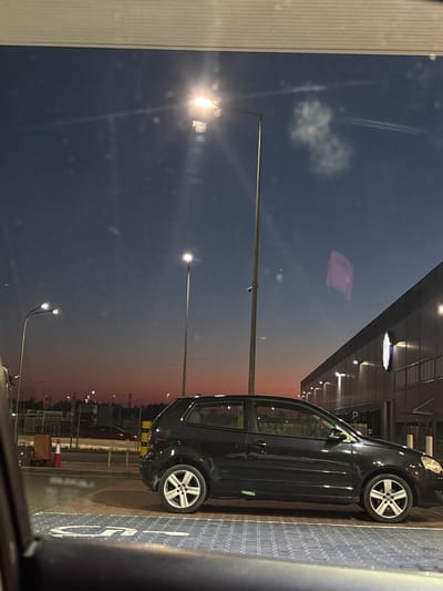 This media file depicts an exterior view of a black car parked on a textured, patterned ground. The car is seen from the side, with its rear wheel and part of its body visible. The car is parked next to a building with a modern facade, featuring a row of lights along its edge and a dark, reflective surface that shows a distorted reflection of the sky and clouds.

The scene is set during twilight, as indicated by the gradient sky transitioning from a deep blue to a warm orange hue near the horizon. The presence of streetlights, one of which is prominently lit and angled towards the sky, further suggests it's evening or early morning. There are faint streaks in the sky that could be contrails from an aircraft. Scattered white specks in the sky might be distant lights or atmospheric particles.

The ground where the car is parked has a distinct pattern, possibly indicating a parking lot or walkway. A white symbol, resembling a wheelchair accessibility icon, is clearly visible on the ground near the car.

In the background, across from the car and building, there are more lights and structures, suggesting an urban or commercial area. The overall mood is calm and somewhat melancholic due to the fading light and the solitary presence of the car. There are no people visible in the scene. The location context is Sękocin Stary, Poland.