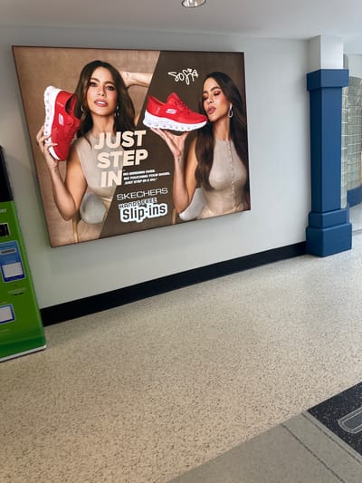 The image shows a Skechers advertisement poster featuring Sofia Vergara in what appears to be an indoor location, possibly a store, based on the presence of a tiled floor and structural elements overhead. The poster highlights Skechers Hands Free Slip-ins shoes, with Sofia Vergara holding a pair of red Skechers sneakers. The poster's text emphasizes the convenience of these shoes, indicating that no bending or touching is required to put them on. There is also a green object under the poster. Location details such as city and street are unavailable from the image.