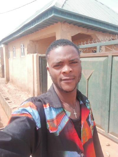 A medium close-up image depicts a male individual, appearing to be in his late 20s or early 30s, posing for a selfie in the city of Bahit, Nigeria. The man has a dark skin tone, short dark hair, and a trimmed beard and mustache. He is wearing a black collared shirt with vibrant, abstract multi-colored patterns in red, blue, orange, and green on the front panels and shoulders. A dark undershirt is visible beneath the open collar. Around his neck, he wears a silver-colored chain with a prominent silver-colored crucifix pendant. He gazes directly at the camera with a neutral expression.
The background consists of a single-story building on the left, constructed from unpainted light brown material, possibly mud or plaster, with visible wear. It features a green corrugated metal roof with an overhang and three white-framed windows on the visible side. To the right, a dark green metal gate with decorative scrollwork along the top edge and an embossed triangular pattern on its lower section is partially visible. The ground is unpaved reddish-brown dirt, with a concrete drainage channel running parallel to the building on the left. The scene is brightly illuminated by direct daylight, under a light, possibly hazy, sky.