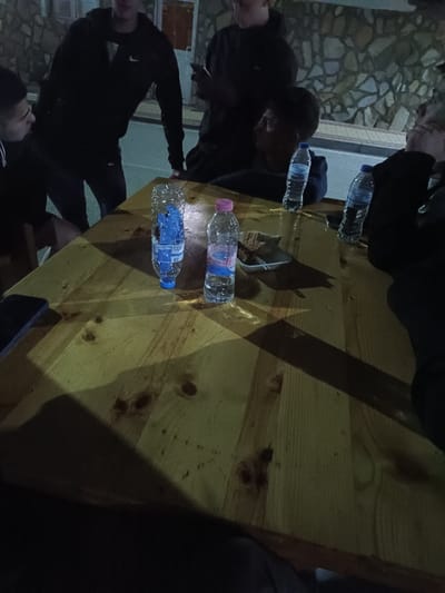A group of people, likely young males, are gathered around a large wooden table outdoors at night. The table has several water bottles and a small dish containing food on it. One person is wearing a Nike sweatshirt. The location is identified as the city of Pripek, Bulgaria based on the water bottle brand. The backdrop features a stone wall and a dimly lit street or pathway. It is a casual social gathering.