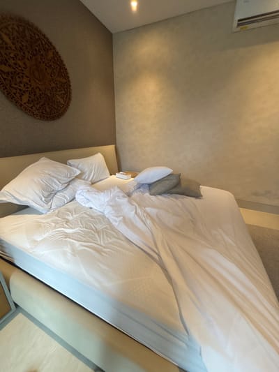 An overhead shot captures a partially unmade bed with white linens. Several pillows are near the head of the bed. A dark brown, oval rug with a complex pattern is visible in the lower left corner, placed on a beige carpet. The walls are light beige with a textured pattern. A window is at the top.
