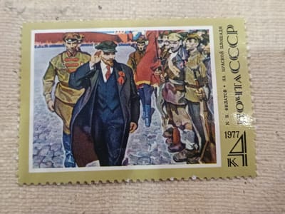 This is a postage stamp from the USSR featuring a depiction of Vladimir Lenin and other figures on Red Square in Moscow. The stamp has a scalloped edge and a pale yellow border. In the center of the stamp, Lenin is shown in a dark suit, white shirt, and red tie, with a red ribbon on his lapel. He is wearing a cap and has his hand raised to his forehead as if saluting or shielding his eyes. He is walking among a group of people in what appear to be military or worker uniforms, some carrying a red flag. The background depicts a stylized urban scene with buildings. To the right of the image, in vertical text, are the words "ПОЧТА СССР" (POSTAGE USSR) and "К. В. ФИЛАТОВ • НА КРАСНОЙ ПЛОЩАДИ" (K. V. FILATOV • ON RED SQUARE). Below this text, the year "1977" is printed, along with a graphic symbol. The stamp itself is not located in Onaldy, Turkmenistan.
