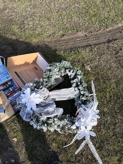 The scene shows a Christmas wreath, decorated with white flowers and silver ribbon, lying on a grassy area adjacent to a muddy track. A partially open cardboard box is positioned next to the wreath. The box contains various DVDs, one of which is titled "Battle of Britain". The location is inferred to be Chirk, United Kingdom, based on the title of the DVD, which references a significant historical event involving the United Kingdom. There are no people visible in the image.