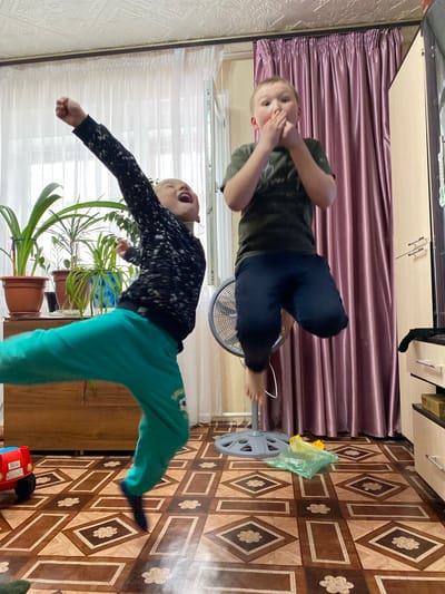 Two boys are in a room in Bely Klyuch, Russia. One boy is lying on a fan wearing a green shirt and navy pants, holding his hands to his face. The other boy is jumping with his right arm raised, wearing a black and white long-sleeved shirt and turquoise pants. There are houseplants in brown pots on a wooden dresser. The floor is brown and patterned. A burgundy curtain hangs on the wall behind the boy on the fan. A red toy car sits on the floor near the jumping boy.
