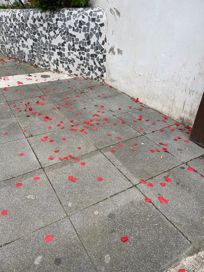 The scene depicts a section of a concrete paved sidewalk in Buñol, Spain. The ground is composed of grey, rectangular concrete paving stones laid in a grid pattern, exhibiting minor stains and wear. Scattered across the entire visible paved area are numerous small, bright red, synthetic or fabric petals, consistent in size and shape, resembling either heart or teardrop forms.
To the left, a low, curved retaining wall or planter box, approximately waist-height, is visible. This wall is adorned with a mosaic composed of irregularly shaped black, various shades of grey, and white tiles. Green foliage is partially visible above the top edge of this mosaic wall.
To the right, a plain, vertical wall, finished in white plaster, occupies the remainder of the background. This white wall shows signs of wear, including dark streaks of dirt or moisture near the base, minor peeling paint, and scuff marks. A dark brown, vertical rectangular object, possibly part of a door frame or a fixture, is partially visible along the extreme right edge of the frame, adjacent to the white wall. No individuals, animals, or direct actions are present within the captured scene. The lighting suggests an outdoor daytime setting.