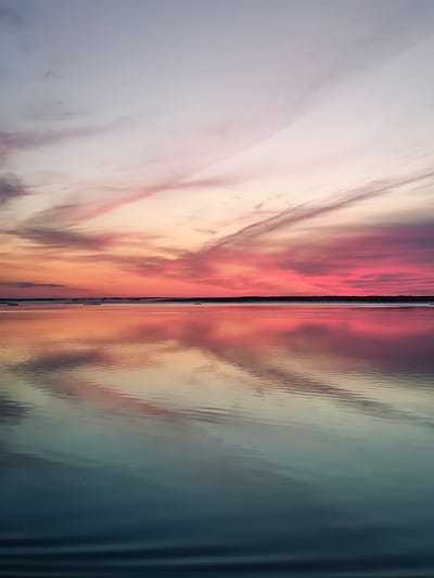 A serene sunset over the water, with vibrant hues of pink, orange, and purple painting the sky. The sky transitions into a pale grey at the top. The colors are reflected beautifully on the calm surface of the water, creating a mirror-like effect. The horizon is marked by a dark line, possibly a treeline or distant landmass, with a few dark shapes visible on the water. The water itself has subtle ripples, adding texture to the reflection. The scene evokes a sense of peace and tranquility. The location is not specified as Chaika, Russia in the image itself.