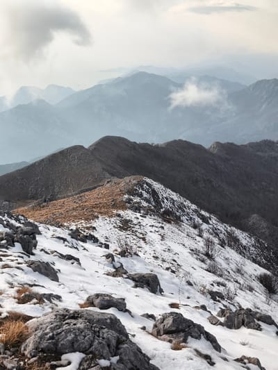 The image displays a panoramic mountainous landscape in Mali Mikulići, Montenegro, under an overcast sky. The immediate foreground features a rugged, snow-dusted slope composed of exposed grey limestone rocks and patches of dry, golden-brown grass. Sparse, bare woody shrubs are scattered across the snow. A prominent, dark mountain ridge runs diagonally through the midground; its nearer face exhibits a mix of snow, rock, and golden-brown vegetation, while its further side is densely covered in snow with scattered bare trees. In the background, multiple successive mountain ranges recede into the distance, appearing increasingly hazy and bluish-grey due to atmospheric perspective. Wisps of white cloud drift among these distant peaks. A faint body of water is discernible on the far left horizon, beyond the layers of mountains. No individuals, animals, or man-made structures are visible.