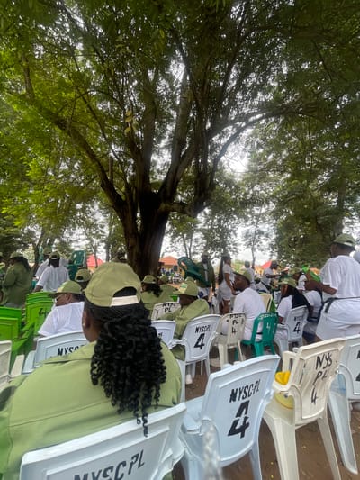 This media file shows a group of people, likely participants in a National Youth Service Corps (NYSC) program, seated outdoors under a large tree. The individuals are predominantly wearing white t-shirts and green caps, which are commonly associated with NYSC activities in Nigeria. The scene appears to be a gathering or ceremony, given the arrangement of chairs and the formal attire.

The setting is outdoors, with a dominant presence of lush green foliage from a large tree overhead, dappling sunlight through its branches. This suggests it's daytime, likely a bright and possibly warm day, given the outdoor gathering. The ground appears to be dirt or earth, typical of an outdoor venue.

Visible text on the white plastic chairs includes "NYSC PL" and a large number "4", which might indicate seating arrangements or participant identification. The people are seated and seem to be attentively facing forward, suggesting they are watching something or listening to an address. Some individuals have their heads bowed, possibly in prayer or deep thought. The overall mood seems to be one of solemnity or focused attention.

The context provided mentions Emure-Ekiti, Nigeria, which aligns with the presence of NYSC participants. The image captures a moment of communal gathering and engagement, possibly related to orientation, a meeting, or a specific event within the NYSC program.