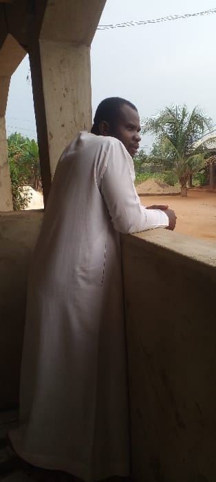 An adult male stands in a shaded, semi-outdoor area in Agunmo, Nigeria, leaning against a concrete balustrade. He is positioned facing right, with his back partially to the camera, his head turned to look towards the right out of frame. The individual wears a long-sleeved, full-length white garment. His forearms rest on the top surface of the unpainted concrete balustrade, with his hands clasped or resting together. A substantial concrete pillar frames the left side of the scene. The background reveals an overcast sky above an unpaved, reddish-brown ground surface. Scattered vegetation, including a prominent palm tree and other green foliage, is visible. A large pile of light-colored material, possibly sand or earth, is present in the mid-ground. Faint utility lines are discernible against the sky.