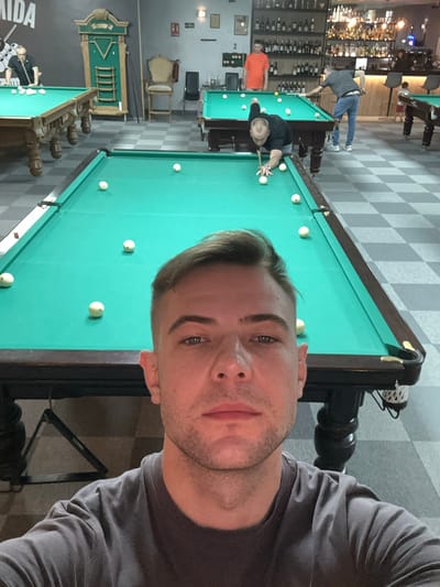 The image depicts a man taking a selfie in what appears to be a billiard hall. In the background, other individuals are engaged in playing billiards at several tables. The hall features a checkered floor, and a bar area is visible, suggesting the location is a recreational establishment. The overall setting points to an indoor entertainment space. There is no explicit indication of its location in San Vicente Del Raspeig/sant Vicent Del Raspeig, Spain.