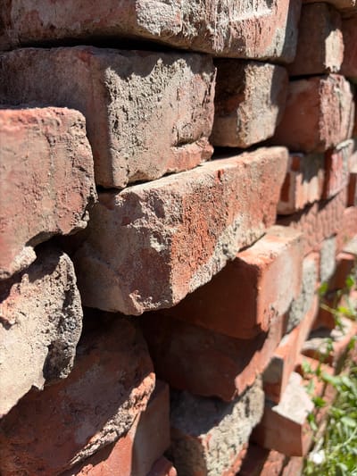 The image is a close-up, low-angle shot of a stack of weathered, reddish-brown bricks. The bricks have a rough, textured surface with visible signs of age and wear, including chipped edges and patches of greyish mortar. Sunlight casts harsh shadows across the surfaces, highlighting the uneven textures. In the lower right corner, out-of-focus green foliage is visible, suggesting the stack is outdoors. The image does not contain any people or visible actions. Based on the provided information, this scene is located in Krugloye, Russia.