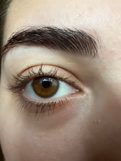 The media file is a close-up, vertically oriented photograph of a person's eye. The eye is open and looking towards the viewer. The iris is a warm brown color, with a dark pupil in the center. Eyelashes frame the eye, appearing dark and somewhat sparse on the upper lid and sparser on the lower lid. A portion of the eyebrow is visible in the upper left of the image; it is dark and well-defined. The skin surrounding the eye is light-toned with some subtle texture and faint marks. The lighting appears to be soft, highlighting the natural contours of the face and the details of the eye. There is no discernible text, weather, or specific environmental context visible in the image itself. The focus is entirely on the eye and immediate surrounding facial features.