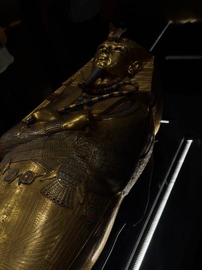 This media file showcases a sarcophagus, likely an ancient Egyptian artifact, made of highly polished gold. The sarcophagus is intricately decorated with detailed carvings and inlaid precious stones in shades of turquoise, red, and blue. A notable feature is the depiction of a human face on the lid, characteristic of pharaonic burial practices. The sarcophagus rests in a dimly lit display setting, with bright artificial lights illuminating it from above, creating dramatic highlights and shadows on the golden surface. The background is predominantly black, emphasizing the artifact. The specific context of Kafrat Nassar, Egypt, suggests this could be an exhibit at a museum or archaeological site in the region, possibly related to a significant discovery or historical figure. No text is visible in the image.