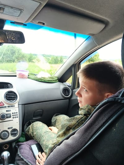 In the foreground of the image, a young boy sits in the passenger seat of a car, looking out the window. He is wearing a camouflage jacket and pants. His hands are resting on his lap, with a smartphone placed on top of his legs. The car's dashboard is visible, with air vents, a radio, and control buttons. In the background, a rural landscape unfolds, with trees and a road stretching into the distance. The sky is bright, suggesting daylight. The scene appears to be set in Medvedevka, Russia, a rural location characterized by its natural surroundings.
