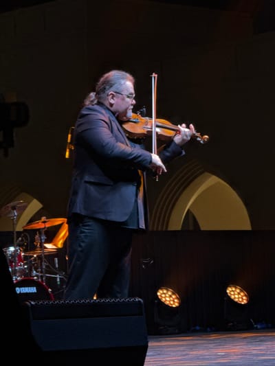 A man in a suit plays a violin. He is facing left with his head tilted back. His hair is long and pulled back. Behind him are stage lights, a sound system, and drums. The stage has a dark background with a tan circular design. The lighting gives the scene a warm tone.