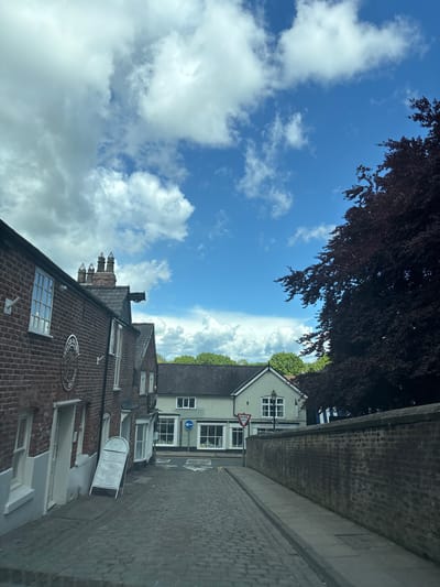 The image shows a street in Knutsford, United Kingdom. The sky is blue with scattered white clouds. A tree with dark red leaves overhangs the scene. On the right side of the street is a tall brick wall, below which is a cobblestone road. Buildings, including a light green building with a "Give Way" sign, are visible along the street. A dark brick building with decorative brickwork and a sign that reads "The Knutsford Tapestry" and an A-frame sign that reads "Watkins Welcome" are also seen.