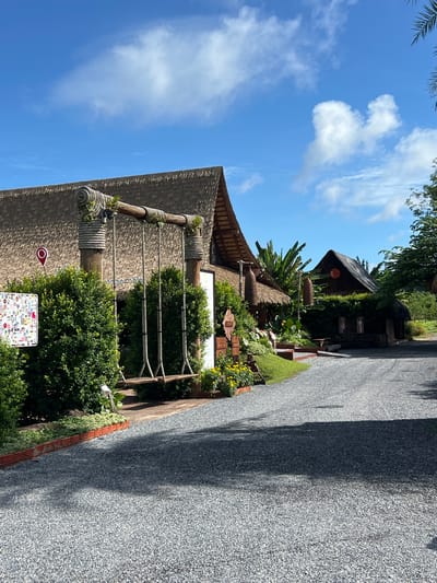 The image depicts a sunny day in Tambon Pa Kae Bo Hin, Thailand, with a clear blue sky dotted with white clouds. The scene features a charming, rustic building with a thatched roof, surrounded by lush greenery and vibrant yellow flowers. A gravel path runs alongside the building, leading into the distance. The architecture suggests a tropical setting, with exposed wooden beams and rope accents.
Visible details include a large, woven cylindrical object hanging from a wooden beam above the building's entrance, possibly a decorative element or a traditional lamp. Several signs are also present: one is a red and white location marker, similar to a map pin, and another is a white board adorned with numerous colorful stickers, hinting at a place of interest or a souvenir shop. The building appears to be a single-story structure, possibly a cafe, restaurant, or guesthouse, given its attractive presentation and landscaping. The overall atmosphere is serene and inviting, conveying a sense of relaxed tropical living.