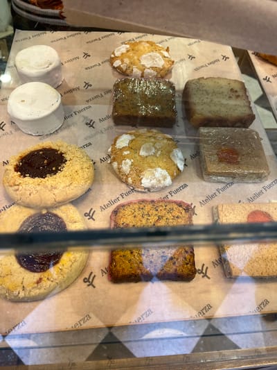 This close-up shot captures a variety of baked goods and pastries displayed in what appears to be a bakery or cafe in Olivos, Argentina. The items are arranged on light brown parchment paper, visible through a glass display case with reflections.
On the top row, from left to right, there is a rectangular loaf or cake speckled with poppy seeds, followed by a square pastry wrapped in plastic, featuring an orange-colored fruit piece on top. Another light-colored rectangular item is partially visible to the far right.
The middle row showcases a round, light-colored cookie or scone with white frosting or sugar, next to a darker rectangular item wrapped in plastic. Another similar round cookie/scone is to its right, and then a reddish-brown, textured rectangular cake, possibly a carrot or fruit cake.
The bottom left of the display holds two small, cylindrical, white items. To their right are two large, round, light-colored cookies with a dark, jam-like fruit filling in their centers.
No people are visible in the image. The parchment paper beneath the items is adorned with a repeating logo and the text "Atelier Fuerza," which is also fragmented into "erza," "A," "elier," and "Fuer" across the different visible sections. The logo appears to be a stylized 'AF' or two overlapping 'f's. The reflections on the glass and a vertical metal frame indicate the items are behind a transparent barrier. The time of day and weather are not discernible.