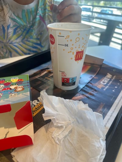 A close-up, eye-level, indoor shot shows a McDonald's cup and a crumpled white napkin on a black tray. The cup, featuring a Coca-Cola logo and orange circular designs, is positioned to the right of the frame, with a visible 0.4L marking. To the left of the cup, a corner of a colorful box with cartoon characters and a red accent is visible. The background is blurred but suggests a restaurant environment with a person wearing a tropical print shirt and sunlight filtering through blinds. The scene is set in Húmera-somosaguas-prado Del Rey, Spain.