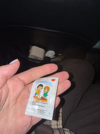 An image captures a person's hand holding a small "Love Is..." cartoon card or wrapper, likely taken inside a vehicle in Kiselevka, Russia. The lighting is dim, suggesting either night-time or an enclosed space.
The primary subject is a human hand, exhibiting white nail polish on the thumb. The hand is holding a rectangular piece of paper or foil, which features a classic "Love Is..." comic strip illustration. The cartoon depicts a boy with a shovel, digging in the ground, while a girl stands beside him, looking on. Above the cartoon, the Russian phrase "Любовь это..." (Love is...) is visible. Below the illustration, text in Cyrillic reads "...договориться похоронить прошлое." (agree to bury the past.), with an English translation directly beneath it: "agree to bury the past." A number "54" is printed in the top right corner of the card. Further down, small text includes manufacturer or distributor details: "Kent Grp", "Cumhuriyet mah. 2253. Sokak No: 11 41450 Gebze-Kocaeli www.cadburyturkey.com Серия 7, 7th Series", indicating a Turkish origin for the product, possibly associated with Cadbury Turkey.
In the blurred background, parts of a dark car interior are visible. To the left, in a dark console or storage compartment, a white rectangular object resembling a power adapter or charger can be seen, along with what appears to be crumpled tissue paper. To the right, a patterned fabric, possibly plaid, is visible, suggesting a blanket or seat cover. The overall scene appears personal and candid.