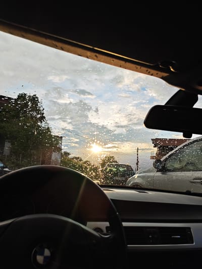 The image shows the view from inside a car, a BMW, looking out through the rain-spattered windshield. The steering wheel is in the foreground, with the BMW logo visible. Beyond the windshield, the scene depicts an urban setting at sunset. A house with a red-tiled roof and a building behind trees, utility poles, and other vehicles are visible. The presence of a Bulgarian license plate 'E 4144 KH' on a dark-colored car outside places the location in Bulgaria. The presence of specific buildings are not discernable, therefore the location in Tserovo cannot be confirmed. The image does not show any people.
