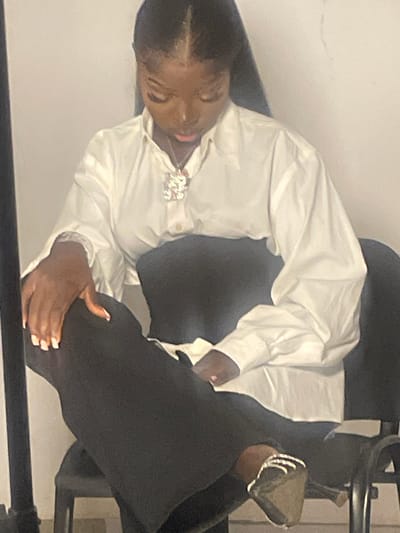 A young Black woman is seated in a chair, facing downwards, with her legs crossed. She is wearing a white button-down shirt with puffed sleeves and a sparkling pendant necklace. Her black pants are wide-legged, and she is wearing high-heeled shoes adorned with rhinestones. The background is a plain, light-colored wall, and a black tripod is partially visible to the left of the frame. The lighting is somewhat dim, creating soft shadows. There is no indication of the specific location within South Ropp, Nigeria, beyond it being an indoor setting with a plain wall.