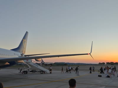 An outdoor, wide-angle photograph captures a scene at an airport on the outskirts of Rimini, Italy, at sunset. The sky transitions from a pale blue at the top to a vibrant orange and yellow near the horizon. In the foreground, a Ryanair airplane is parked on the tarmac, its tail fin visible with the airline's logo. A mobile boarding staircase is attached to the aircraft, and several people are disembarking or walking around it. Further in the background, other aircraft are visible, including a TRANSAVIA plane, and various airport support vehicles and structures. A group of people, appearing to be passengers and airline staff, are scattered across the tarmac, some walking with luggage, others standing and talking. The ground is paved with asphalt, marked with yellow lines. The overall atmosphere suggests the end of a flight and the process of deplaning.