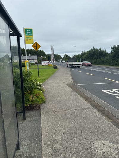 The image depicts a street scene in Stradone, Ireland, on a cloudy day. A bus stop shelter with a glass enclosure is visible on the left side of the frame. Adjacent to the shelter is a bus stop sign indicating "Stop 131641" with route numbers 109, 109x, and 170. A yellow information post stands near the bus stop sign. A paved sidewalk runs alongside the road, bordered by a strip of grass with flowering plants and bushes. The road itself has clear lane markings and traffic signs. Several vehicles, including a white utility truck and a red car, are driving on the road. In the background, houses and dense foliage are visible. A large banner with text and an image of a person, likely an advertisement, is also present on the right side of the image. The overall impression is a typical roadside scene in a suburban or rural area.