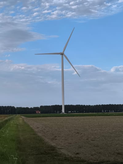 This image shows a lone wind turbine standing tall in a rural landscape. The turbine, a clean white structure, has three blades, one of which is angled upwards, pointing towards the bright blue sky. Fluffy white clouds dot the sky, adding depth and texture to the scene. The turbine is situated in a field of dry, tilled soil, with a strip of vibrant green grass running alongside a dirt path to the left. In the background, a dense line of dark green trees forms a horizon, with a hint of red-roofed buildings peeking through the foliage. The overall impression is one of serene, open country, with the wind turbine symbolizing a modern element within a natural setting. The location is specified as Wieringerwerf, Netherlands.