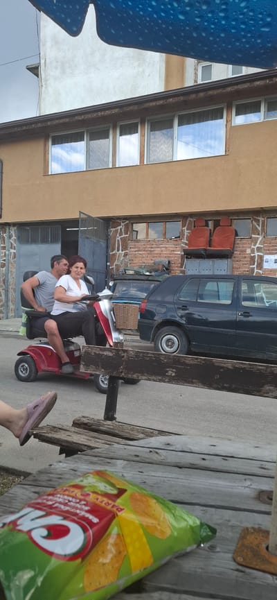 The image, captured in Dobrinishte, Bulgaria, features a man and a woman seated together on a red mobility scooter in an outdoor setting during daytime. The woman, with reddish-brown hair and a white t-shirt, sits behind the man, who wears a grey t-shirt. Both are smiling brightly at each other, indicating a joyful and casual interaction.

The background shows two distinct buildings. On the left, a light brown stucco building with multiple windows reflects a cloudy sky. In the center, a brick building with a stone-clad lower section features smaller windows and a large metal garage door. Stacked red car seats are visible against the brick wall. A black van is parked behind the scooter, and another car with a brown wicker basket on its back is partially visible further back.

In the foreground on the right, part of a rustic wooden bench or table is visible. A green, red, and yellow bag of chips rests on it, displaying the words "ON" and "PARTY". A person's foot, wearing a pink slipper, is also visible, suggesting another individual is present at the table. To the far left, a blue, textured object with visible water droplets partially frames the scene. The ground beneath the scooter and vehicles is a grey paved surface.