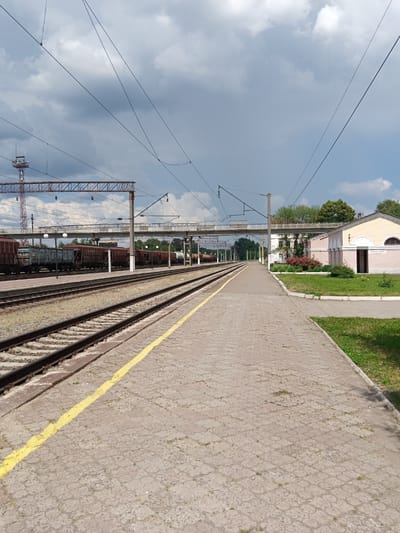 The image depicts a view of a train station platform in Romodan, Ukraine. In the foreground, a paved walkway with a yellow line runs parallel to a set of train tracks. The walkway appears to be made of interlocking concrete pavers. To the left of the walkway are multiple train tracks, with freight cars visible on some of them. In the mid-ground, a pedestrian overpass spans the tracks, supported by concrete pillars. A light-colored building, possibly a station building or public facility, is situated to the right of the walkway and overpass, with architectural details including arched windows and a double door. The building is pink and yellow. Lush green grass and red flowering bushes are visible in front of the building. The sky is cloudy with patches of blue. Overhead, complex electrical wires for the trains are suspended from poles and structures. A tall communication tower with antennas is visible in the distance on the left. The scene suggests a functional, operational train station in a somewhat rural or suburban setting. There are no people visible in the frame.